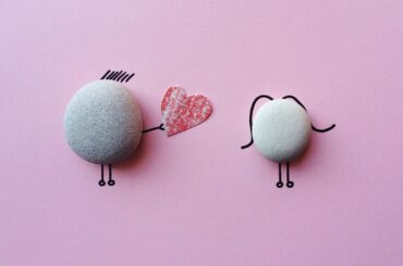 dos piedras redondeadas con extremidades dibujadas a mano. Una figura le entrega un corazón de papel rojo a la otra sobre un fondo rosa.
